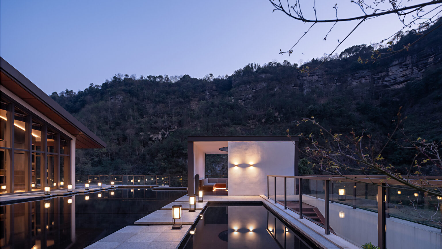 Rooftop terrace at dusk with soft lighting, framed by a modern building on the left and a cozy seating area on the right. Surrounded by a forested mountain backdrop.