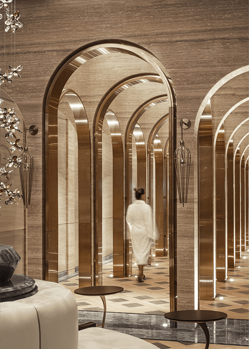 Luxurious resort spa interior with arched golden doorways and marble walls. A person in a white robe walks through the opulent, serene hallway.