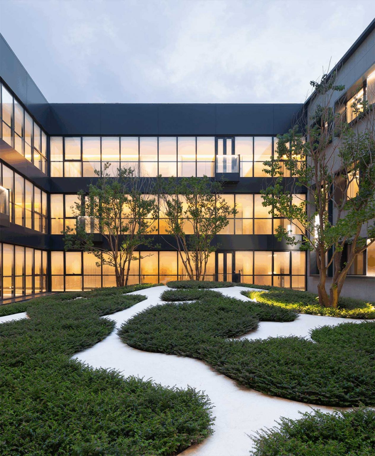 Modern hotel building courtyard with geometric shrub patterns on white gravel. Warmly lit windows line the three-story structure, creating a serene evening ambiance.