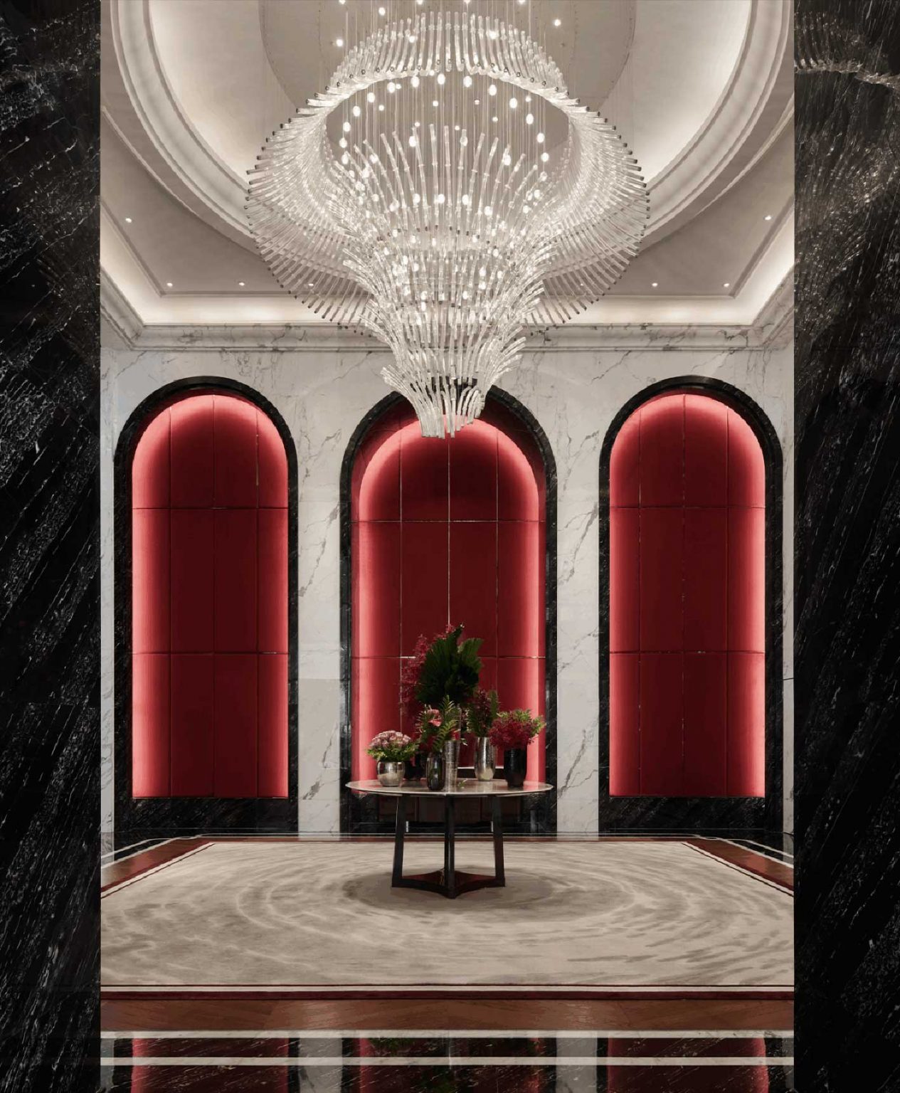 A luxurious hotel lobby featuring a grand chandelier, elegant arched design elements, and rich red accents, complemented by a central table adorned with flowers, creating a sophisticated and welcoming ambiance.