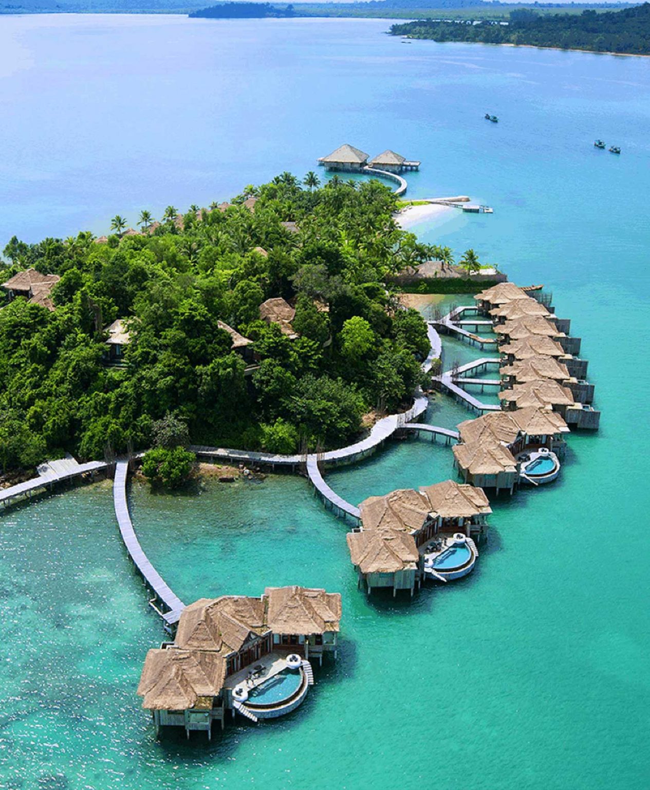 An aerial view of an idyllic tropical resort shows thatched-roof bungalows on stilts, connected by winding wooden walkways over a clear turquoise ocean surrounding a lush green island.