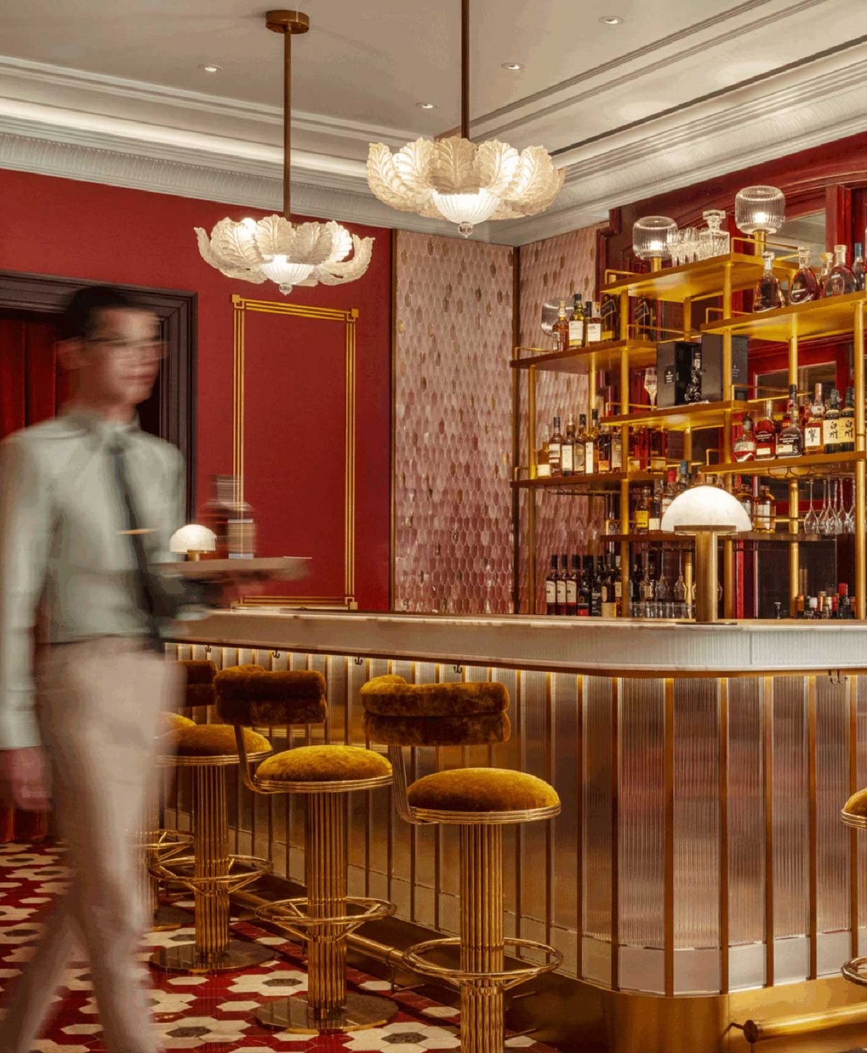 Sophisticated private club bar with velvet stools, gold accents, and intricate lighting. A server in motion enhances the elegant, lively ambiance.